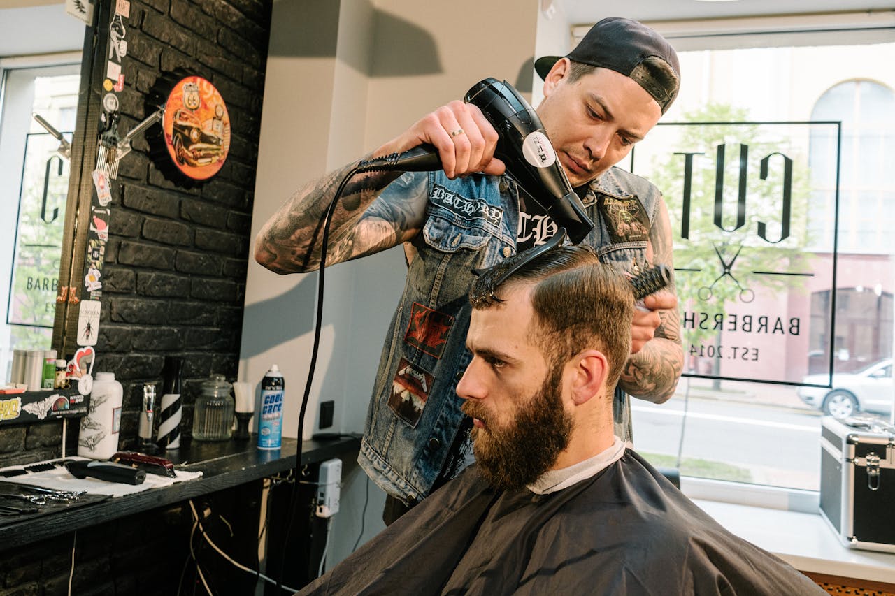 A barber uses a blow dryer to style a client's hair in a trendy barbershop setting.