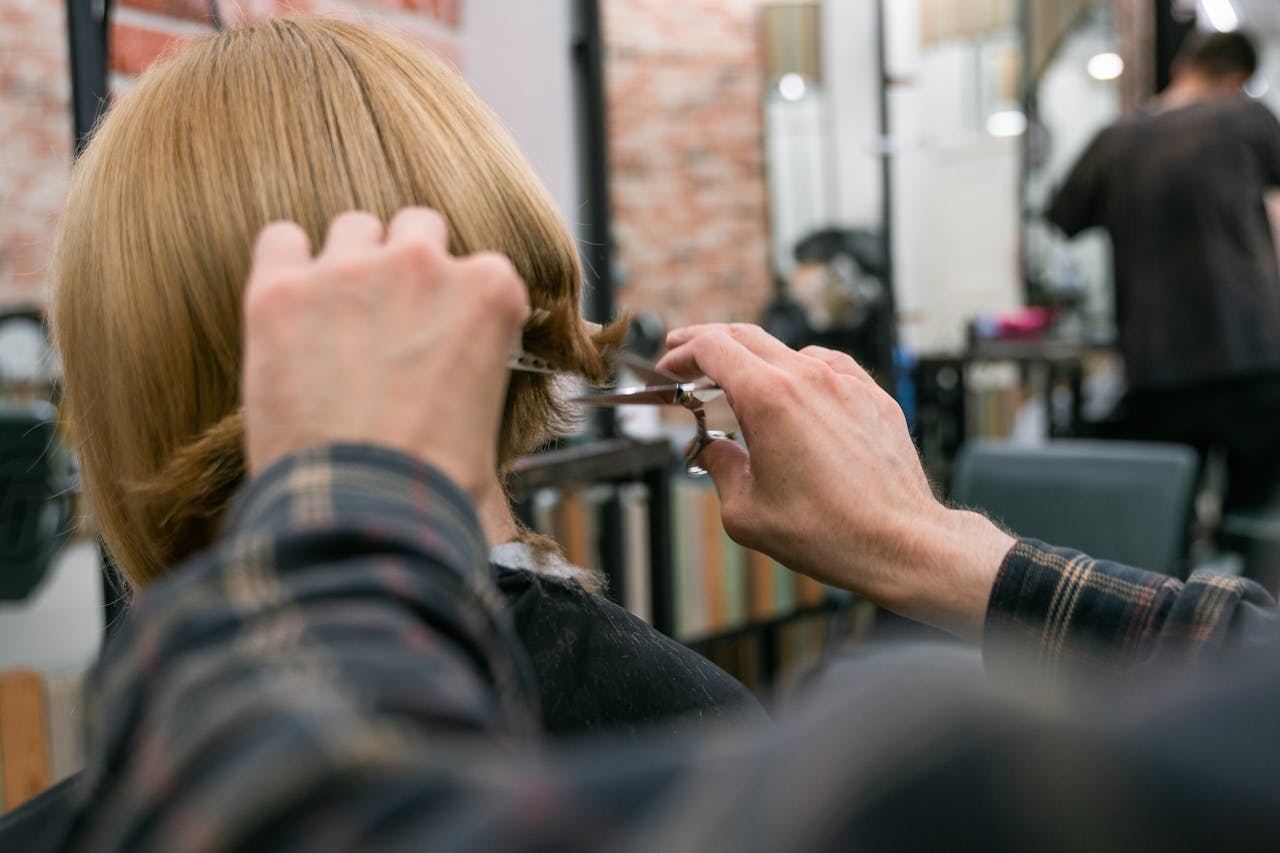 Hairdresser cutting woman's blonde hair in salon with scissors and mirror.