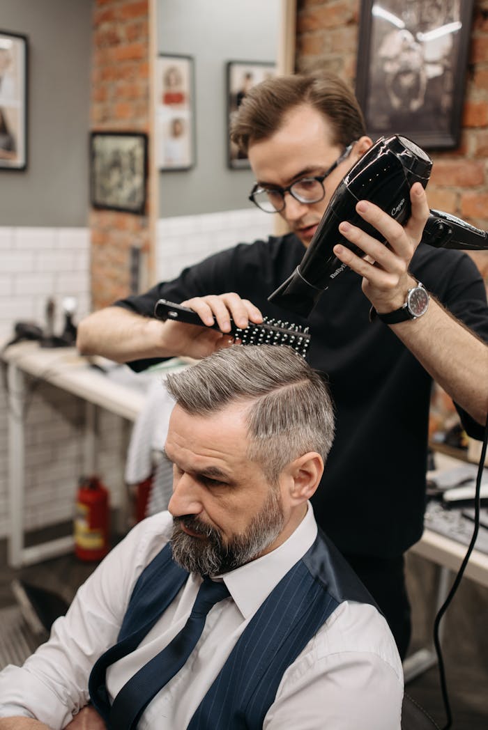 Barber styling a bearded client's hair using a blow dryer and brush in a trendy salon.